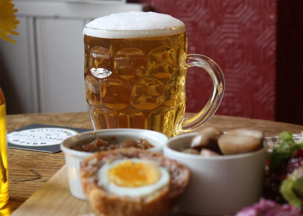 A pint of golden ale with a frothy head served in a traditional dimpled glass tankard, resting on a wooden pub table beside a Butcombe Bitter coaster. In the foreground, a halved Scotch egg with a soft golden yolk is accompanied by a ramekin of caramelised onion chutney and a ramekin of pickled onions. A side of mixed leafy salad is partially visible, with a red patterned wallpaper adding warmth to the cosy pub setting.