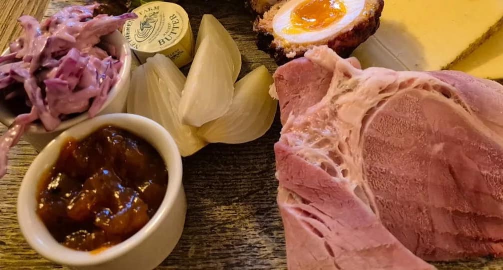 Close-up of a traditional British Ploughman’s Lunch on a rustic wooden board. The image features thick slices of Wiltshire-style ham, a wedge of mature cheddar cheese, half a Scotch egg with a bright orange yolk, peeled pickled onions, creamy red cabbage coleslaw in a white ramekin, a pot of dark fruity chutney, and a pat of Wyke Farms butter. The textures and colours highlight the hearty, homemade feel of this classic cold pub meal.