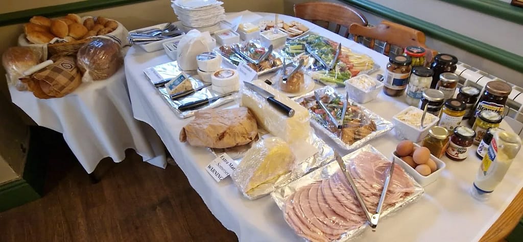 This image features a well-organized buffet spread, likely intended for a "Ploughman's Lunch" or a similar event showcasing local foods. Here's a detailed description:
### Bread Selection:
- On a small round table to the left, there's a basket filled with an assortment of bread rolls, loaves, and slices. The loaves include crusty artisan styles, dark rye, and soft white bread. Knives are placed nearby for easy serving.
### Cheese Spread:
- The main table prominently displays a variety of cheeses. There are round wheels of soft cheese (e.g., Brie or Camembert), wedges of hard cheese (such as cheddar), and some cheeses wrapped in wax or foil for freshness.
### Deli Meats and Protein:
- To the lower-right side of the table are trays of sliced ham, beautifully arranged in overlapping rows. A bowl of boiled eggs in their shells is also included, adding a traditional and protein-rich element.
### Salad and Vegetables:
- At the centre, trays of fresh vegetables like cherry tomatoes, celery, cucumber, and mixed greens are neatly organized. Some trays contain sandwiches or wraps, perhaps as a nod to a lighter option within the Ploughman's theme.
### Condiments and Pickles:
- A variety of jars are arranged along the back edge of the table, containing chutneys, pickled onions, Branston pickle, pickled eggs, and mustard. These are quintessential accompaniments to a Ploughman’s-style spread.
### Other Highlights:
- Butter dishes and spreads are positioned near the bread and cheese.
- There’s also a "WARNING" sign near one of the cheese items, potentially indicating a strong or particularly distinctive cheese, like Stilton or one made with unpasteurized milk.
### Presentation:
- The table is covered in a white cloth, creating a clean and inviting backdrop. The food is served on foil-covered trays and small bowls, with serving tongs, knives, and spoons for hygiene and convenience.
This setup emphasizes traditional British fare, celebrating simplicity, freshness, and variety.