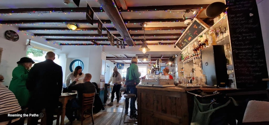 A cozy interior view of the bar area at the Three Mariners in Tenby. The room features exposed wooden beams on the ceiling, decorated with colorful string lights, creating a warm and inviting atmosphere. Several patrons are seated at wooden tables, enjoying drinks and conversation, while others stand near the bar, giving the space a lively feel. The bar itself, made of rustic wood, is stocked with various bottles and glasses, while a chalkboard menu hangs on the right side, listing available drinks or specials. The scene captures a charming, bustling pub environment.