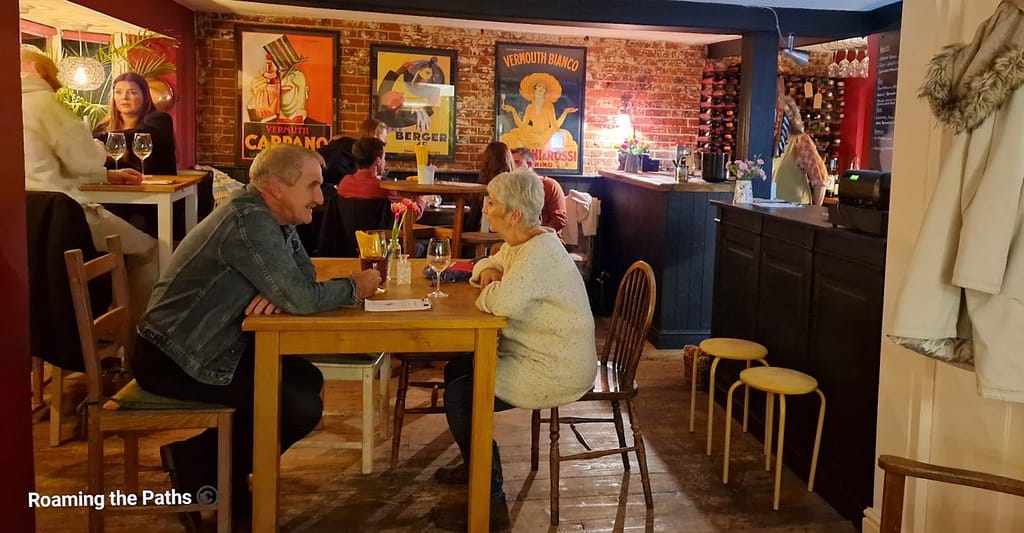This image shows a cozy, intimate scene at Alfred's Wine Bar, featuring guests seated around simple wooden tables in a relaxed atmosphere. The exposed brick walls are adorned with vintage posters, enhancing the warm, rustic decor. In the background, a small bar area is visible with neatly arranged wine bottles on display. People are engaged in conversation, with glasses of wine on the table, contributing to a laid-back, friendly ambiance. The lighting is soft and inviting, creating a comfortable setting for enjoying drinks and company.