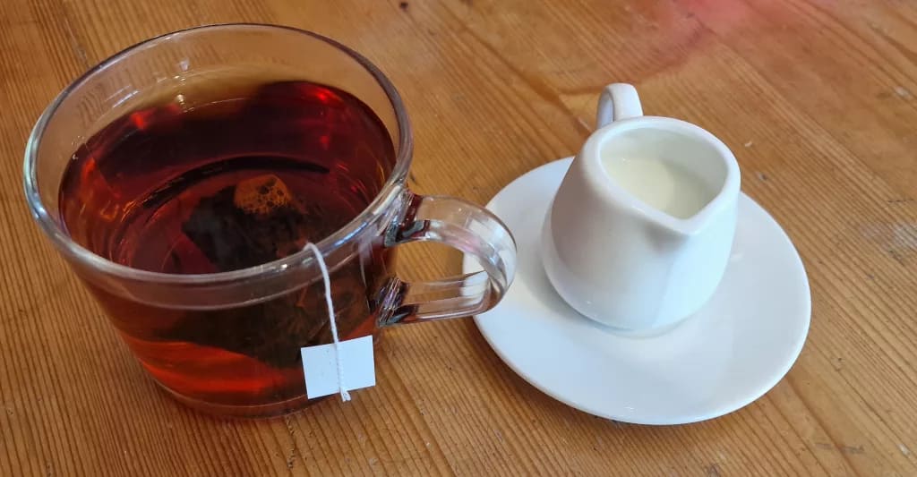 A clear glass mug of black tea with a teabag string hanging over the side, placed on a wooden table next to a small white jug of milk on a saucer.