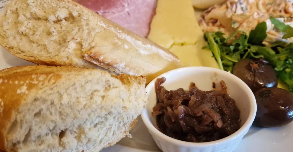Close-up of a Ploughman’s lunch showing torn slices of crusty bread, a small white ramekin of onion chutney, glossy pickled onions, fresh salad leaves, cheddar cheese, and a slice of ham in the background.
