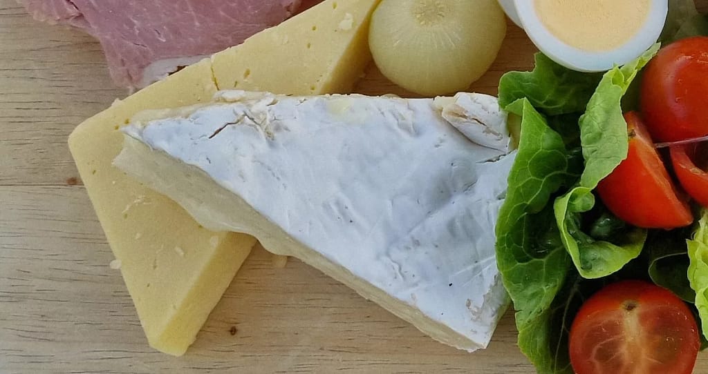 Close-up of a wedge of creamy brie with a white rind resting atop a thick slice of mature cheddar cheese on a wooden platter. Surrounding the cheeses are a pickled onion, half a boiled egg, fresh green lettuce leaves, and vibrant cherry tomatoes, offering a colourful and appetising arrangement.