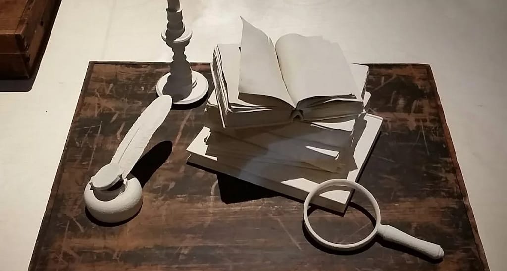 A still life arrangement of monochrome white objects on a dark wooden surface, featuring a stack of open and closed books, a candlestick, a quill in an inkwell, and a magnifying glass. The objects are styled to appear sculptural and uniform in colour, creating a visually striking contrast with the worn, textured table beneath them.