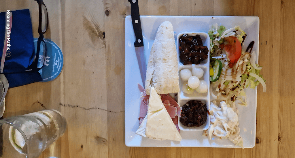 A flatlay view of a Ploughman’s lunch served on a square white plate. The meal includes a wedge of cheddar cheese and a baguette positioned diagonally, along with sliced ham. The center of the plate features a divided dish containing pickled onions, dark chutney, and cucumber slices. On the right side, there is a fresh salad of lettuce, tomato, cucumber, and onion drizzled with balsamic glaze, next to a creamy coleslaw. A knife rests alongside the baguette, and glasses and a pair of eyeglasses are visible in the background on a wooden table.
