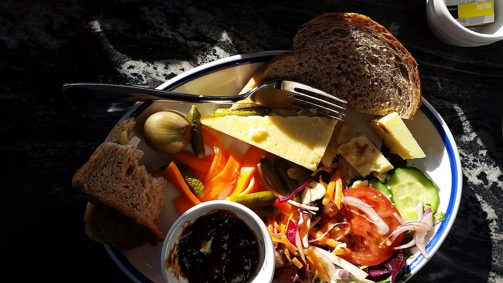 The image shows a flat view of a Ploughman’s lunch, served on a plate from "Salt and Malt." The meal includes:
A wedge of cheddar cheese.
Slices of hearty wholegrain bread.
Pickled vegetables, including a whole pickled onion and gherkins.
Carrot sticks alongside the pickles.
A fresh salad with sliced cucumber, tomato, red onion, and shredded vegetables.
A small dish of dark chutney or relish.
The plate is bathed in sunlight, casting warm highlights on the fresh, colourful ingredients, creating an inviting, rustic feel.