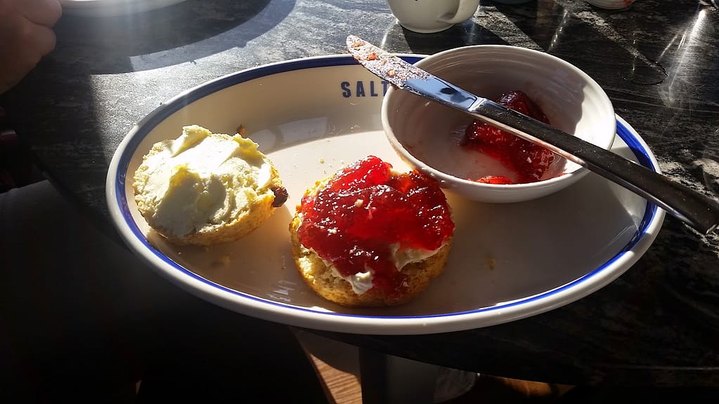 
The image shows a classic cream tea setup, likely from a café. On a plate labeled "Salt and Malt," there are two halves of a scone. One half is topped with a generous amount of clotted cream, and the other with both clotted cream and a thick layer of bright red strawberry jam. Next to the scone is a small bowl containing more jam, with a knife resting across it. Sunlight is streaming across the table, highlighting the rich textures of the scone and toppings, creating a warm and inviting scene for an afternoon tea.