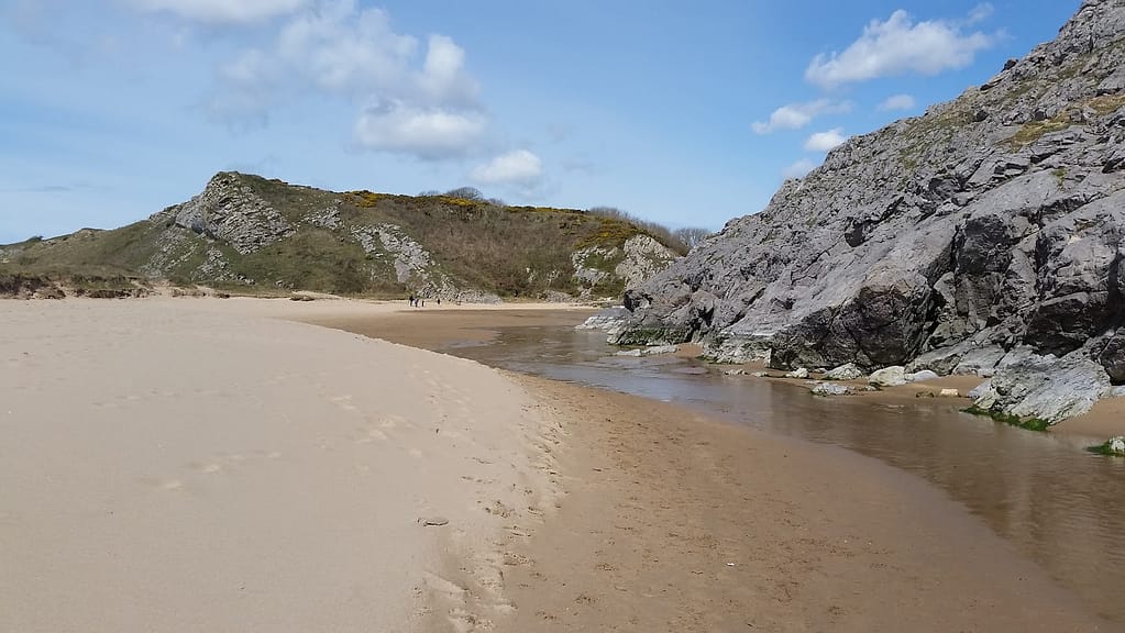 The image shows a scenic beach scene, likely taken at Bosherton Broad Haven Beach. The foreground is a wide expanse of sandy shore with gentle footprints leading toward the distance. A small stream or water channel runs alongside the sand, bordered by rugged rock formations on the right side. In the background, there are rolling grassy hills with patches of rocks and shrubs. A few people can be seen walking in the distance, adding a sense of scale to the vastness of the beach. The sky is blue with scattered white clouds, suggesting a clear, pleasant day at the beach.