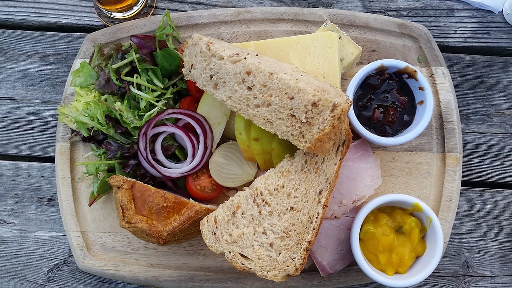 A flat-lay of a Ploughman's lunch at the Mayfly in Fullerton. The meal is served on a wooden board, featuring thick slices of multigrain bread, cheddar cheese, and generous portions of ham. A mini pork pie adds to the hearty presentation, with accompaniments of Branston pickle and bright yellow mustard piccalilli in separate ramekins. A fresh salad of mixed greens, sliced red onions, cherry tomatoes, and pickled onions completes the dish. The rustic wooden table and natural light enhance the inviting, countryside feel of the meal.