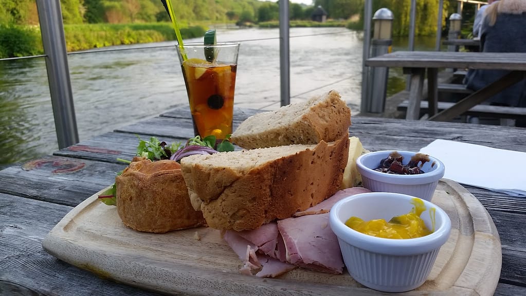 A scenic riverside Ploughman's lunch at the Mayfly in Fullerton. The plate features thick slices of multigrain bread, ham, cheddar cheese, a mini pork pie, and small ramekins filled with Branston pickle and mustard piccalilli. There's a fresh side salad with mixed greens and red onion, complementing the savory elements of the dish. In the background, a glass of refreshing Pimm's with fruit and a cucumber garnish sits on a rustic wooden table, with the serene river flowing nearby, creating a relaxing countryside dining experience.