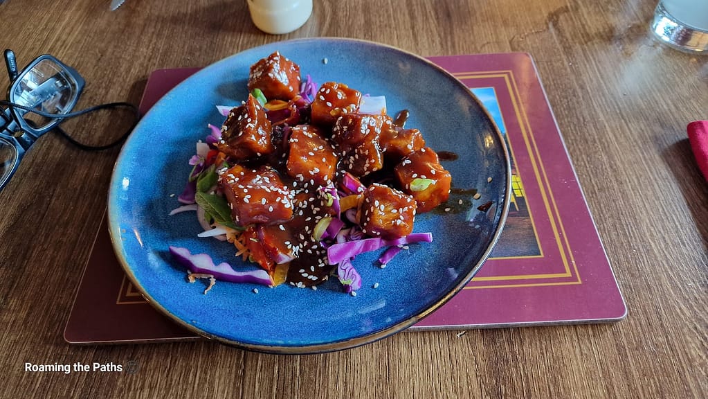 A plate of crispy pork bites coated in a hoisin sauce, sprinkled with sesame seeds, and served on a bed of colorful slaw made of red cabbage, carrots, and green onions. The dish is presented on a blue plate placed on a wooden table, with a pair of glasses and other table items visible in the background.