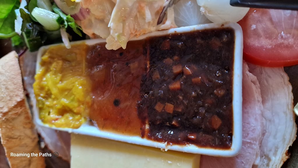 
The image appears to depict a trio of condiments on a tray, likely used as accompaniments for a traditional ploughman's lunch. The condiments include what looks like piccalilli (a yellow, mustard-based pickle with cauliflower or similar vegetables), mango chutney (a sweet and tangy orange sauce), and ploughman’s pickle (a dark, thick chutney with visible vegetable chunks). They are placed on a plate with slices of ham, cheese, and some fresh vegetables like lettuce and tomato.