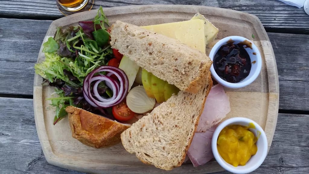 Ploughman’s lunch served on a wooden board: thick slices of brown bread, cheddar cheese, ham, pork pie, salad with onion and tomato, apple slices, pickled onions, chutney, and mustard pickle