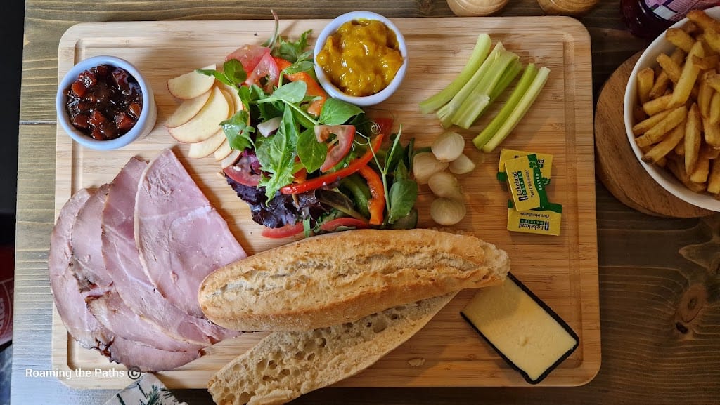 The image is a top-down view of a classic Ploughman’s lunch served on a wooden board at The Woolpack in Sopley. On the left side of the board, several slices of thick, tender ham are arranged neatly. To the left of the ham, a small white dish filled with dark, chunky chutney adds a tangy, sweet element to the meal. In the center of the board, a fresh and colorful mixed salad features vibrant greens, sliced red bell peppers, and juicy tomato wedges. Thin slices of crisp red and green apples are fanned out beside the salad, adding a refreshing crunch. Adjacent to the salad, a small dish contains a bright yellow mustard pickle relish for extra flavor. To the right of the salad, a few sticks of fresh celery and a cluster of small pickled onions offer a sharp, crunchy accompaniment. Two neatly wrapped butter pats sit beside them. The bottom of the board features a golden, crusty baguette, split open to reveal a soft, airy interior. To the right of the baguette, a thick wedge of mature cheddar cheese, pale and crumbly, waits to be paired with the bread and ham. Off to the side, a bowl of golden, crispy French fries adds an indulgent touch to the meal, making this a hearty and satisfying pub lunch. The wooden board and rustic table setting complete the traditional pub atmosphere, while personal items like glasses and condiments scattered in the background hint at a cozy, leisurely meal.