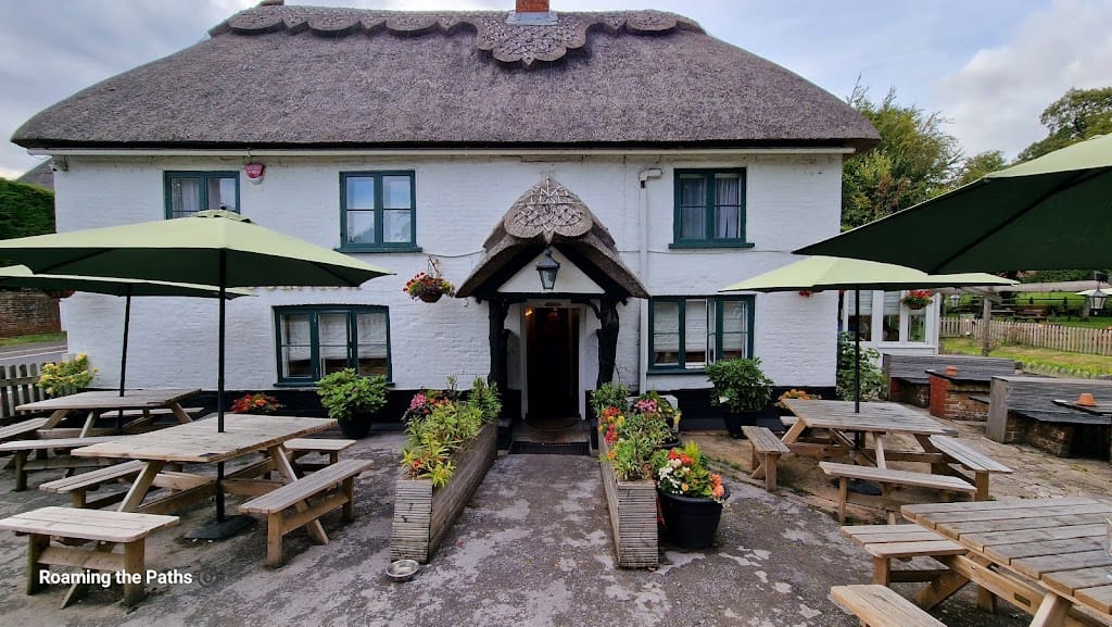 The image shows the front exterior of The Woolpack in Sopley, a charming traditional English pub with a white-painted facade and a distinctive thatched roof. The pub features a quaint, rustic entrance, framed by a decorative wooden arch. Bright flower arrangements in pots and planters line the walkway, adding vibrant color to the scene.
Two large green umbrellas cover wooden picnic benches set outside the pub, creating a welcoming outdoor seating area for patrons. The building's simple design, small windows with dark green frames, and wooden beams give it a cozy, countryside atmosphere. The patio area appears inviting and well-maintained, perfect for enjoying a casual meal or drink.