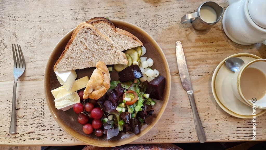 This image shows a beautifully presented ploughman’s lunch on a round brown plate. In the upper section of the plate, three thick slices of rustic whole grain bread are stacked. To the left of the bread, there are generous slices of creamy Brie cheese, and a mini pork pie with golden pastry is nestled nearby. The bottom left features a small bunch of vibrant red grapes, while to the right, a mixed salad of fresh greens, cherry tomatoes, and chopped spring onions sits alongside cubes of pickled beetroot. Tucked next to the bread are sliced pickles and a pile of small, shiny white pickled onions. A teapot, creamer, and cup with saucer sit to the side on a wooden table, ready for tea. A fork and butter knife are placed neatly alongside the plate.