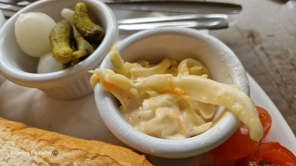 The image shows a close-up view of two small ramekins on a plate, each containing classic ploughman's accompaniments. The ramekin on the left holds a few tiny pickled onions and small gherkins (cornichons), offering a crunchy and tangy bite. The ramekin on the right contains a creamy serving of freshly made coleslaw, with visible strands of cabbage and carrot coated in mayonnaise. In the background, part of a crusty baguette and halved cherry tomatoes are also visible, along with cutlery, suggesting a traditional British ploughman's lunch setting. The textures and colors emphasize the variety of flavors on the plate.