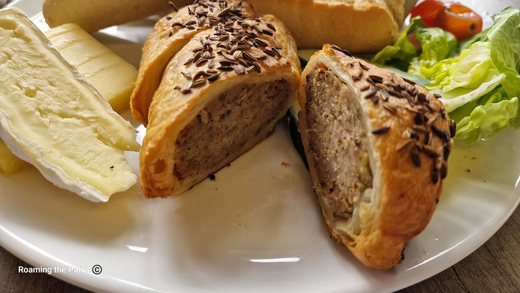 The image shows a close-up of a savory sausage roll, cut in half to reveal its seasoned meat filling. The sausage roll is wrapped in flaky golden pastry and topped with caraway seeds, which add a subtle aromatic crunch. Next to it, there are slices of creamy Brie cheese and blocks of butter, providing a rich and smooth contrast to the sausage roll. In the background, part of a crusty baguette and fresh salad greens, including lettuce and cherry tomatoes, are visible, completing a traditional British ploughman’s lunch. The textures, from the crispy pastry to the soft cheese, create a visually appealing and appetizing dish