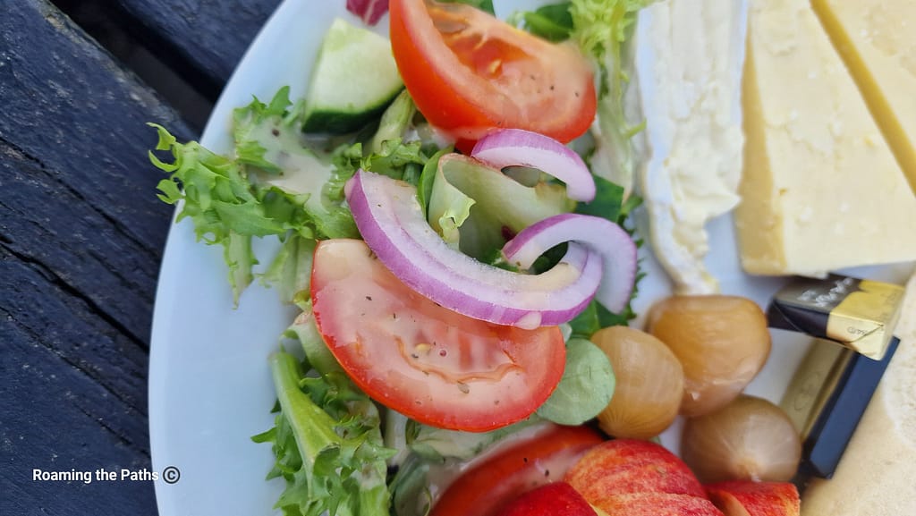 This image showcases a close-up of a fresh, vibrant salad from a Ploughman's lunch. The salad consists of crisp lettuce leaves, thick slices of juicy red tomatoes, and rings of sharp red onion. Accompanying these are chunks of cool cucumber, providing a refreshing crunch. The salad is lightly dressed with a vinaigrette, visible on the leaves and tomatoes, adding a slight sheen to the fresh produce.

To the right of the salad, there are small pickled onions, offering a tangy contrast to the fresh ingredients. The edge of the plate also features wedges of creamy Brie and mature cheddar, alongside wrapped butter pats. The colors of the salad — from the deep reds of the tomatoes and onions to the pale green of the lettuce — are set against the neutral tones of the cheese, creating a beautifully balanced, appetizing presentation. The salad appears fresh, crisp, and perfectly complements the rest of the meal. The rustic wooden table underneath adds a charming, countryside feel to the overall scene.
