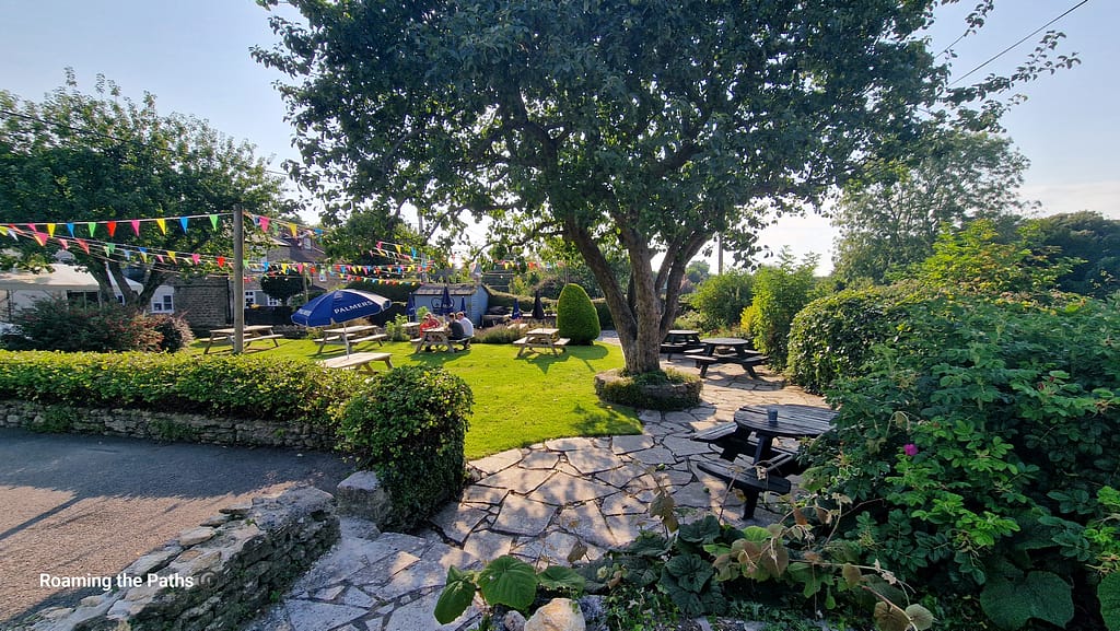 The image depicts a charming beer garden at The Crown Inn, Puncknowle, Dorset. The garden features picnic tables set on lush, well-maintained grass, offering a peaceful and inviting outdoor dining space. Colorful bunting strung across the garden adds a festive touch. A large tree provides shade, and hedges and bushes surround the seating area, enhancing the natural beauty. In the background, the pub's building and Palmers-branded umbrellas can be seen. The tranquil setting is perfect for enjoying a drink or meal, surrounded by greenery and the picturesque Dorset countryside.