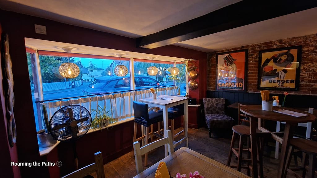 The image shows the cozy interior of Alfred's Wine Bar. The room features a warm, rustic design with exposed brick walls, dark red paint, and wooden beams. A large window dominates one side, framed with simple white curtains and hanging lantern-style lights. There's a mix of seating options, including a tall table with stools and a round wooden table with chairs. A fan, some potted plants, and decorative wall plates add to the ambiance. Vintage-style posters adorn the walls, enhancing the laid-back, welcoming vibe of the space.