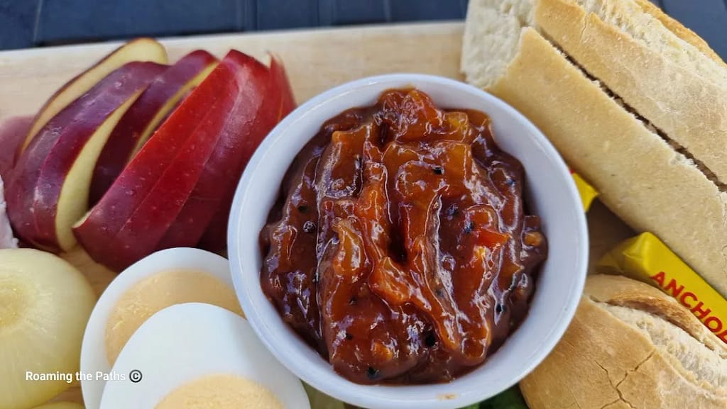 Close-up of a white ramekin filled with chunky, rich brown ploughman’s chutney, featuring visible pieces of onion and spices. Surrounding the chutney are slices of red apple, a halved boiled egg, a pickled onion, and crusty baguette slices with butter packets tucked beside them. The image captures the vibrant, rustic texture and colour of the chutney.