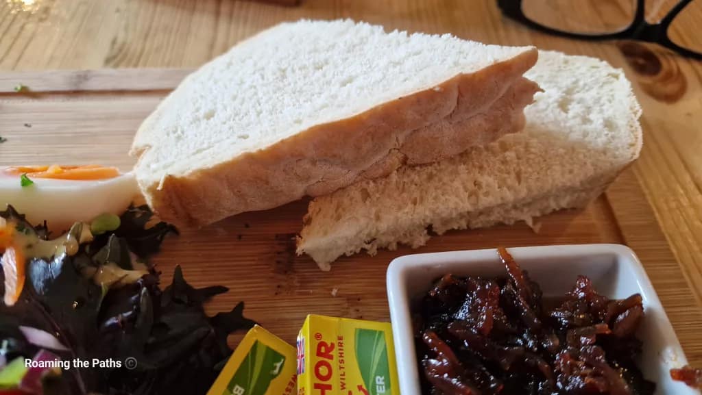 Two thick slices of soft white farmhouse bread on a wooden board, served with butter portions, salad, and a dish of caramelised onion chutney as part of a traditional Ploughman’s lunch