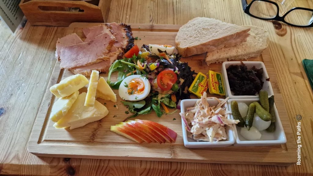 A traditional Ploughman’s lunch served on a wooden board, featuring thick slices of ham, chunks of cheddar cheese, a halved boiled egg on a mixed salad, sliced apple, coleslaw, pickled onions, gherkins, caramelised chutney, packets of Anchor butter, and two slices of white bread. A pair of glasses rests on the table beside the board