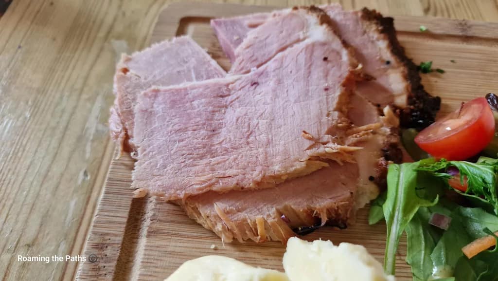 Close-up of thick-cut slices of traditional Wiltshire ham with a peppered edge, served on a wooden board alongside fresh salad and cheese