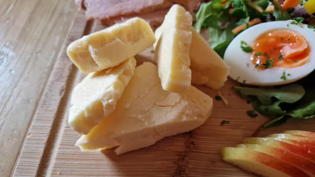 Close-up of chunky slices of crumbly cheddar cheese next to a soft-boiled egg with a runny yolk, served on a wooden board with mixed salad and sliced apple