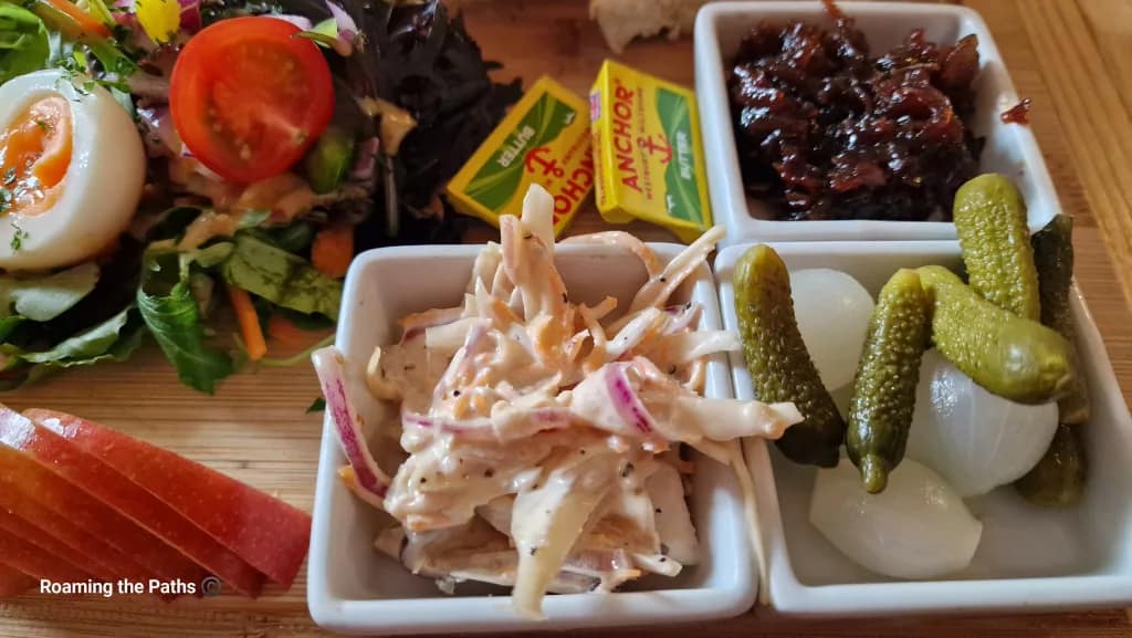 Close-up of a Ploughman’s lunch showing three ceramic dishes containing creamy coleslaw, sweet caramelised onion chutney, and a mix of pickled gherkins and silverskin onions. Nearby are sliced red apple, mixed salad leaves, a halved boiled egg, and mini packs of Anchor butter.