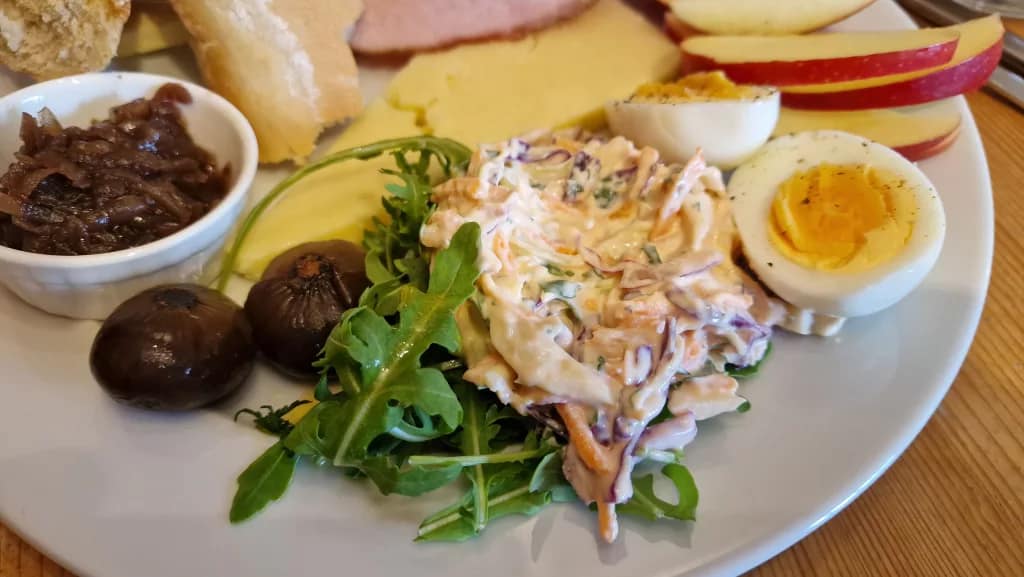 Close-up of a Ploughman’s lunch plate showing creamy coleslaw with shredded carrot and cabbage, fresh rocket leaves, two glossy pickled onions, a small dish of onion chutney, slices of cheddar cheese, and a hard-boiled egg cut in half beside wedges of red apple.