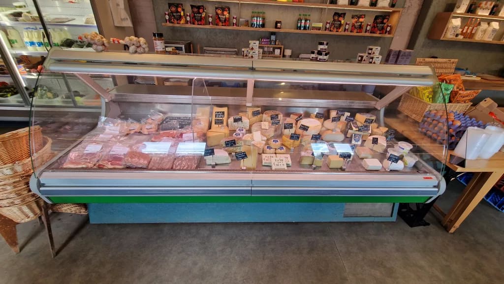 Deli counter in a farm shop displaying a wide selection of cheeses labelled with small signs, alongside cuts of meat. Shelves in the background hold jars, bottles, and packaged goods, with trays of fresh eggs stacked to the side.
