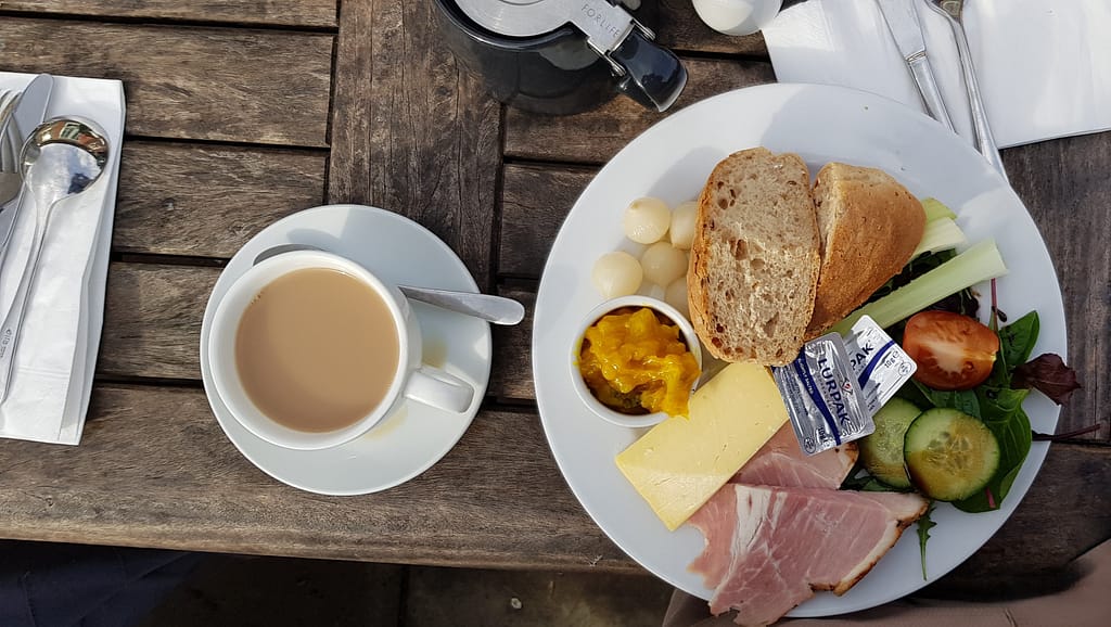 The image shows a flat-lay of a classic Ploughman’s lunch on a rustic wooden table. The plate features a variety of traditional items, including thick slices of ham, a wedge of cheddar cheese, and crusty whole-grain bread. Accompanying the main components are pickled onions, a small pot of vibrant yellow piccalilli, a salad with fresh cucumber, lettuce, and a halved cherry tomato, as well as celery sticks. Two packets of Lurpak butter sit on top of the cheese, ready to be spread on the bread. Next to the plate is a cup of tea with milk, placed on a saucer with a small spoon, and a black teapot with a silver lid. A set of silverware, wrapped in a napkin, lies beside the meal. The setting is casual and outdoors, with natural sunlight illuminating the scene, creating a cozy, relaxed dining atmosphere.