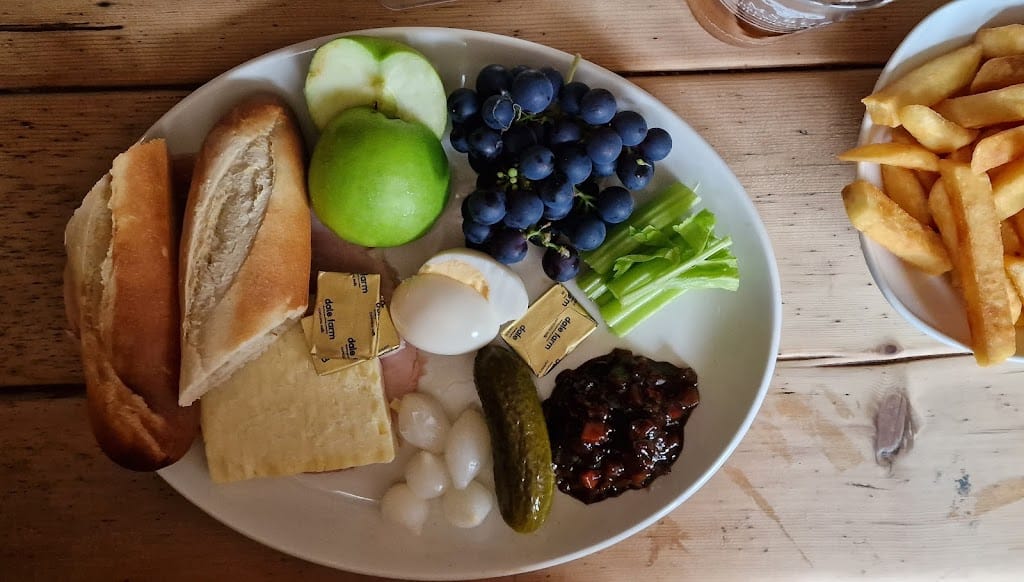 This image features a flatlay view of a traditional ploughman’s lunch, served on a large white plate atop a wooden table. The meal includes a variety of items: two fresh crusty baguette slices and a generous portion of crumbly cheddar cheese. Alongside the cheese, there is a serving of sliced ham, a whole boiled egg, and small pickled onions, all neatly arranged. A large gherkin and a dark chutney are present for added flavor. The plate also includes fresh fruits—a green apple, halved, and a bunch of black grapes—as well as celery sticks for crunch. Small butter packets are placed near the bread and cheese. Off to the side, a small bowl of golden French fries adds a warm, savory element to the meal. The rustic wooden table and the pint of beer in the background hint at a casual pub setting, making this a hearty, visually pleasing ploughman’s lunch.