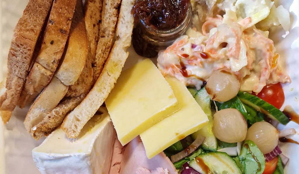 A close-up of a traditional Ploughman’s Lunch featuring thick slices of crusty wholegrain bread, creamy Brie, mature Cheddar cheese, and slices of ham. The plate also includes pickled onions, a small jar of tangy chutney, crunchy salad with cucumber, cherry tomatoes and red onion, creamy coleslaw, and herby potato salad. A hearty, rustic British meal served cold with fresh and colourful ingredients.