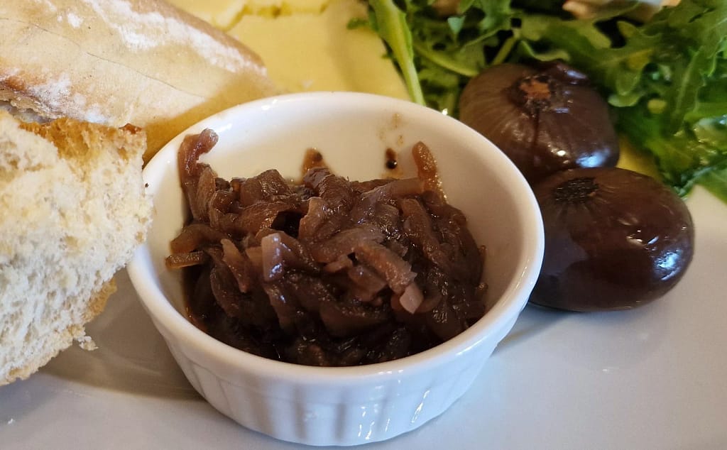 A close-up of a Ploughman’s lunch showing a small white ramekin filled with dark, glossy onion chutney, its soft caramelised strands piled thickly inside. Beside it sit two shiny brown pickled onions, their skins intact and glistening. A torn piece of crusty bread rests to the left, while the edge of fresh green salad leaves and a wedge of pale cheddar can be seen in the background, giving a sense of the hearty platter beyond the frame.