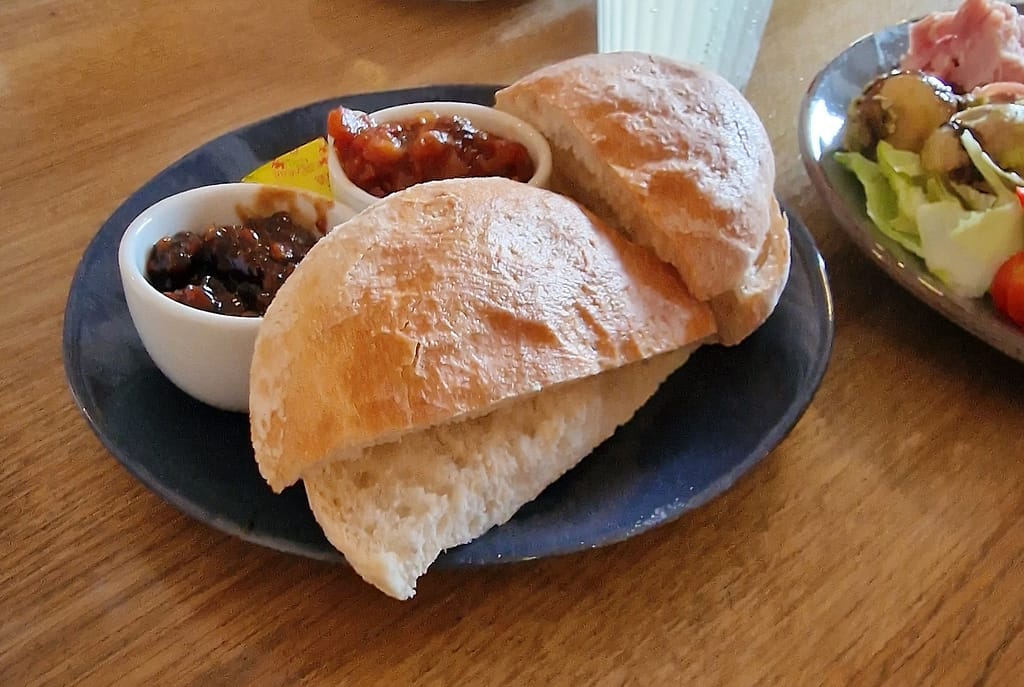 A close-up view of a plate featuring two freshly baked bread rolls, halved and placed on a dark blue ceramic plate. The bread looks soft and fluffy on the inside with a lightly golden, crusty exterior. Accompanying the bread are two small white ramekins filled with condiments. The left ramekin contains a rich, dark chutney, likely a traditional ploughman’s pickle with visible chunks of vegetables. The right ramekin holds a vibrant, reddish chutney with a slightly glossy finish, suggesting a fruity or tomato-based flavor. A small packet of Welsh butter is placed behind the condiments, ready to be spread on the bread. The plate is positioned on a rustic wooden table, adding to the cozy, countryside feel of the meal. In the background, part of a larger plate featuring a fresh salad and slices of ham and cheese can be seen, hinting at a full traditional British Ploughman's lunch.