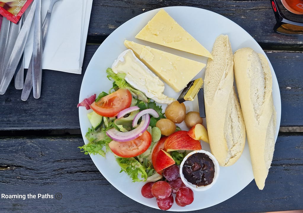 
The image shows a traditional Ploughman’s lunch flatlay, elegantly arranged on a white plate set against a weathered wooden surface. The meal includes two thick slices of mature cheddar cheese and a wedge of soft, creamy Brie. A portion of crusty baguette is sliced lengthwise into two pieces, placed to the right of the cheeses.

On the left side of the plate is a fresh, vibrant salad consisting of crisp lettuce, juicy slices of tomato, thin rings of red onion, and cucumber, all lightly dressed. Nestled beside the salad are several small pickled onions, adding a tangy contrast to the dish.

In the lower part of the plate, fresh fruit provides a refreshing sweetness, with red apple slices and a handful of red grapes. A small ramekin of chutney accompanies the fruit, likely intended as a complement to the cheeses.

Also on the plate are two small packets of butter, suggesting the baguette can be enjoyed with a spread of creamy butter to enhance the overall flavor. The presentation is simple yet appetizing, evoking the rustic charm of an English countryside meal.

Off to the side, silver cutlery wrapped in napkins and a red-tinted object (possibly sunglasses) are visible, hinting at an outdoor dining experience. The entire composition conveys a hearty, fresh, and delicious Ploughman’s lunch perfect for a sunny day.