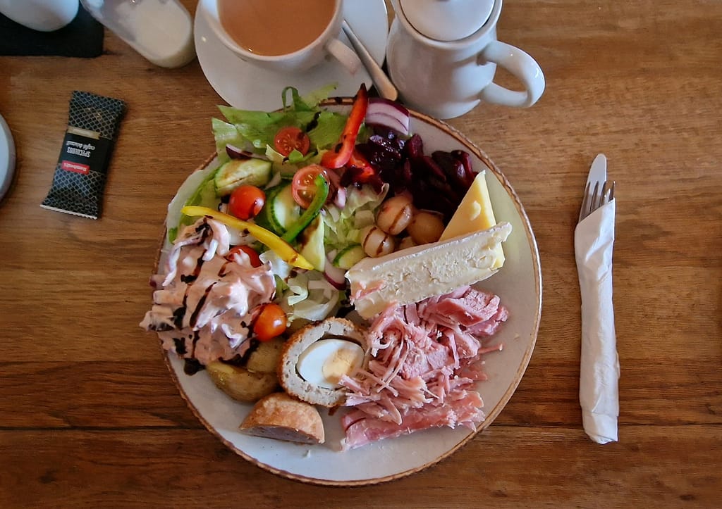A flatlay of a generously portioned Ploughman's lunch served on a round plate. The meal includes a variety of classic components: thick slices of creamy Brie and mature Cheddar cheese, a pile of tender, shredded ham, and half a Scotch egg with its golden yolk exposed. Alongside the proteins, there are pickled onions, sliced beetroot, and a fresh salad with lettuce, cherry tomatoes, cucumber, red onion, and bell peppers, drizzled with a balsamic glaze. The salad also includes a creamy homemade coleslaw. In the foreground, a piece of pork pie is visible, adding a hearty, savory element. The plate sits on a wooden table, next to a neatly wrapped set of utensils and a cup of tea with milk. A teapot and milk jug are placed nearby, enhancing the traditional British meal setting.
