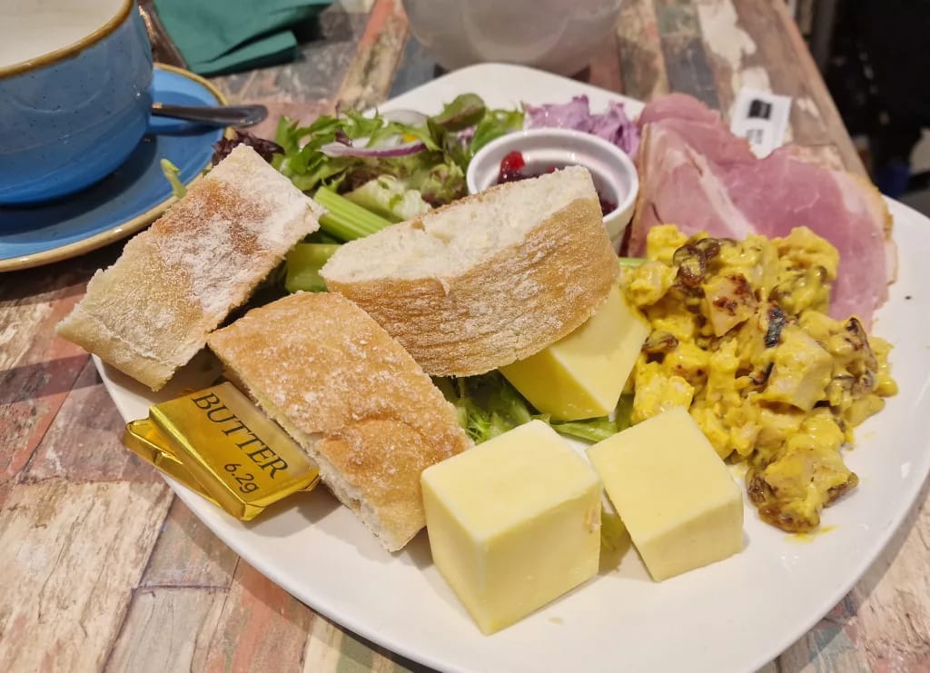 Ploughman’s-style lunch with crusty bread, cubes of cheese, sliced ham, coronation chicken, celery, salad leaves and a ramekin of chutney on a rustic table.