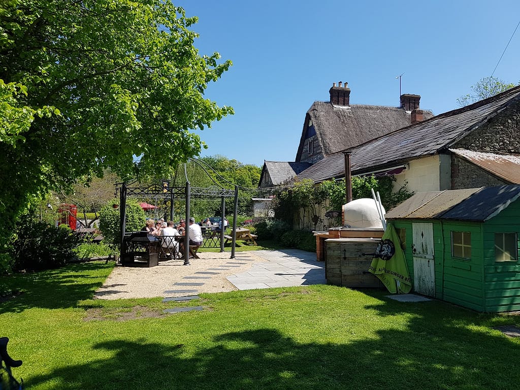 A peaceful outdoor dining area at The Golden Swan, Wilcot, featuring a shaded seating space under a metal pergola. The surrounding garden is lush with greenery, and a traditional thatched roof building sits in the background. The scene captures a sunny day with guests enjoying their time outside amidst natural beauty. A small green shed and outdoor wood-fired oven add to the rustic charm of the setting.