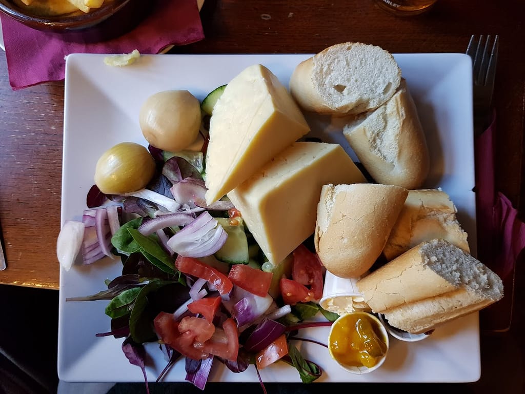 A flat-lay view of a Ploughman’s lunch served at the Boat Inn in Cromford. The meal is arranged on a large white square plate, featuring generous wedges of cheddar cheese, two pickled onions, fresh baguette slices, and a side salad with leafy greens, diced tomatoes, cucumbers, and red onion. Small pots of chutney and mustard add to the meal, with the bread and cheese providing the main focus. The plate is vibrant with colorful vegetables and a well-balanced presentation of classic Ploughman's elements. The rustic table and cozy pub atmosphere add to the charm.