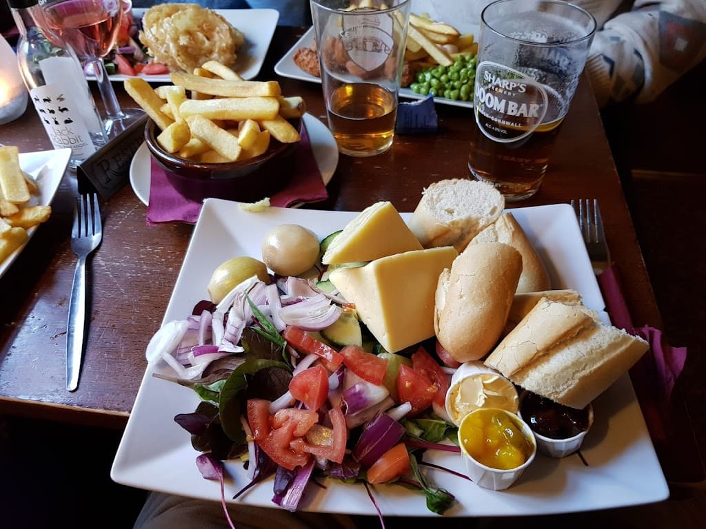 A classic Ploughman's lunch served at The Boat Inn, featuring a rustic presentation on a wooden table. The meal includes thick slices of cheddar cheese and ham, accompanied by fresh mixed salad greens, sliced tomatoes, cucumber, and red onion. A dollop of creamy coleslaw and a portion of pickled onions add traditional elements to the plate. The dish also includes a slice of crusty bread and a small dish of chutney, completing the hearty, quintessential British pub meal. The presentation is simple yet inviting, with natural lighting highlighting the freshness of the ingredients.