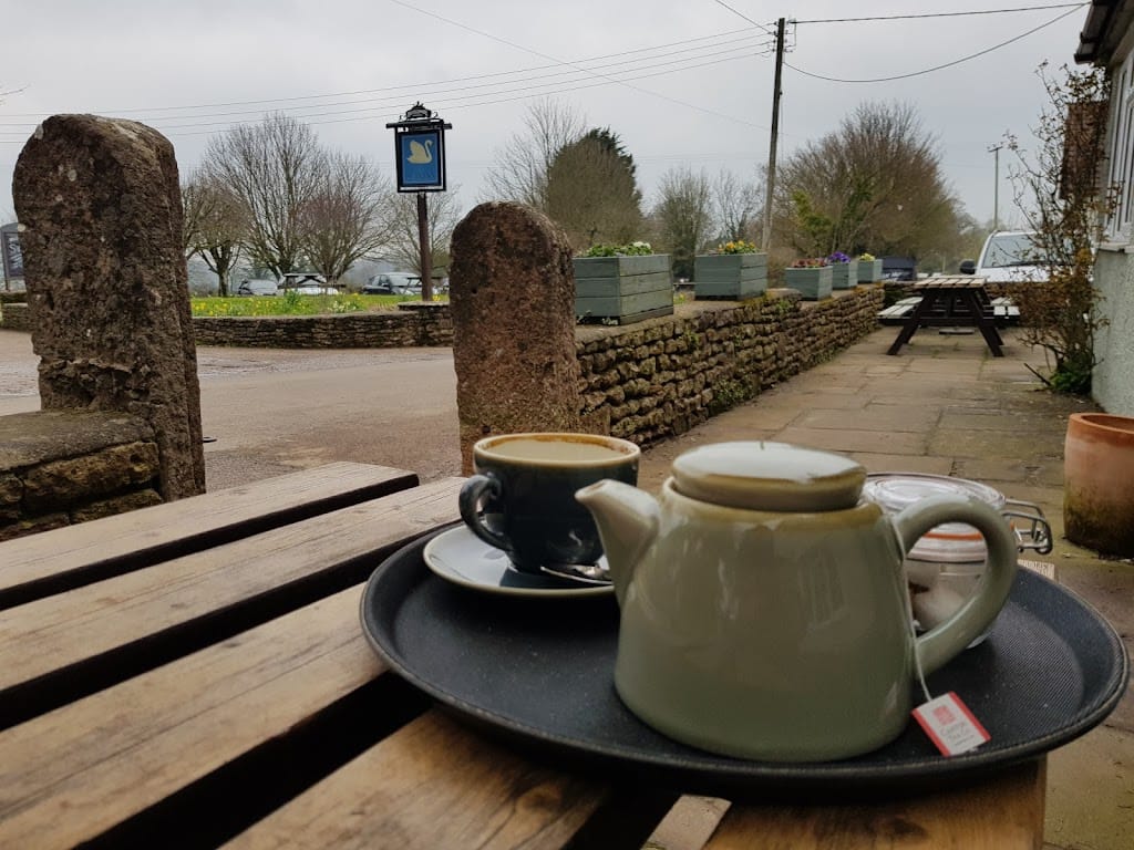 A peaceful outdoor setting at The Swan Inn, Rowberrow, showcasing a tray with a teapot and a cup of tea on a rustic wooden table. The tea is served in a light green ceramic teapot with a matching teacup and saucer, placed beside a small milk jug and a glass jar. The background reveals a stone wall with planters filled with flowers, and the inn’s sign hanging on a post in the distance. A quiet village road and parked cars are visible under a cloudy sky, enhancing the calm and tranquil atmosphere. The scene reflects a charming countryside ambiance perfect for a quiet tea break.