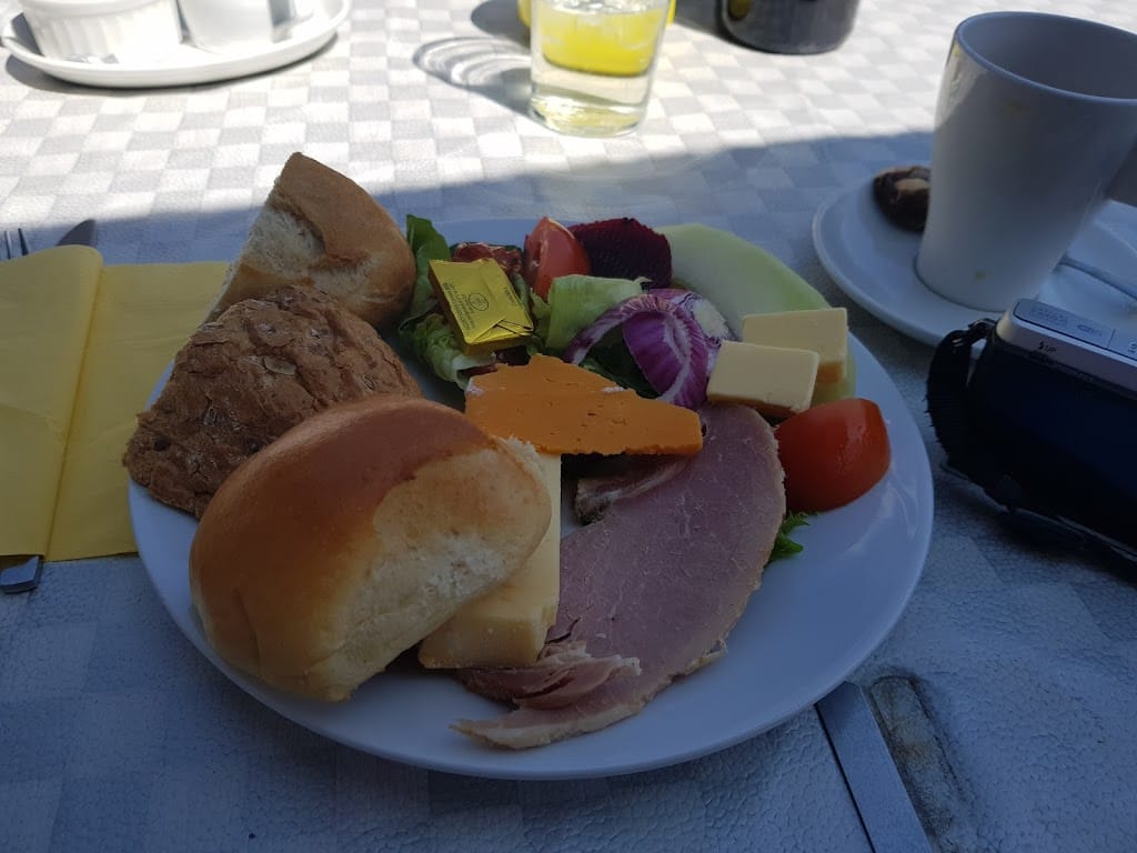A Ploughman's lunch served outdoors on a white plate, featuring two types of bread rolls, slices of ham, cheddar cheese, and a wedge of soft cheese. The plate includes a colorful salad with tomato, red onion, beetroot, and lettuce. A small pat of butter rests on top of the salad along with some cubes of cheese. The setting includes a yellow napkin, a cup of tea, and a camera, suggesting a casual and relaxing meal experience. Sunlight casts soft shadows, enhancing the outdoor atmosphere.