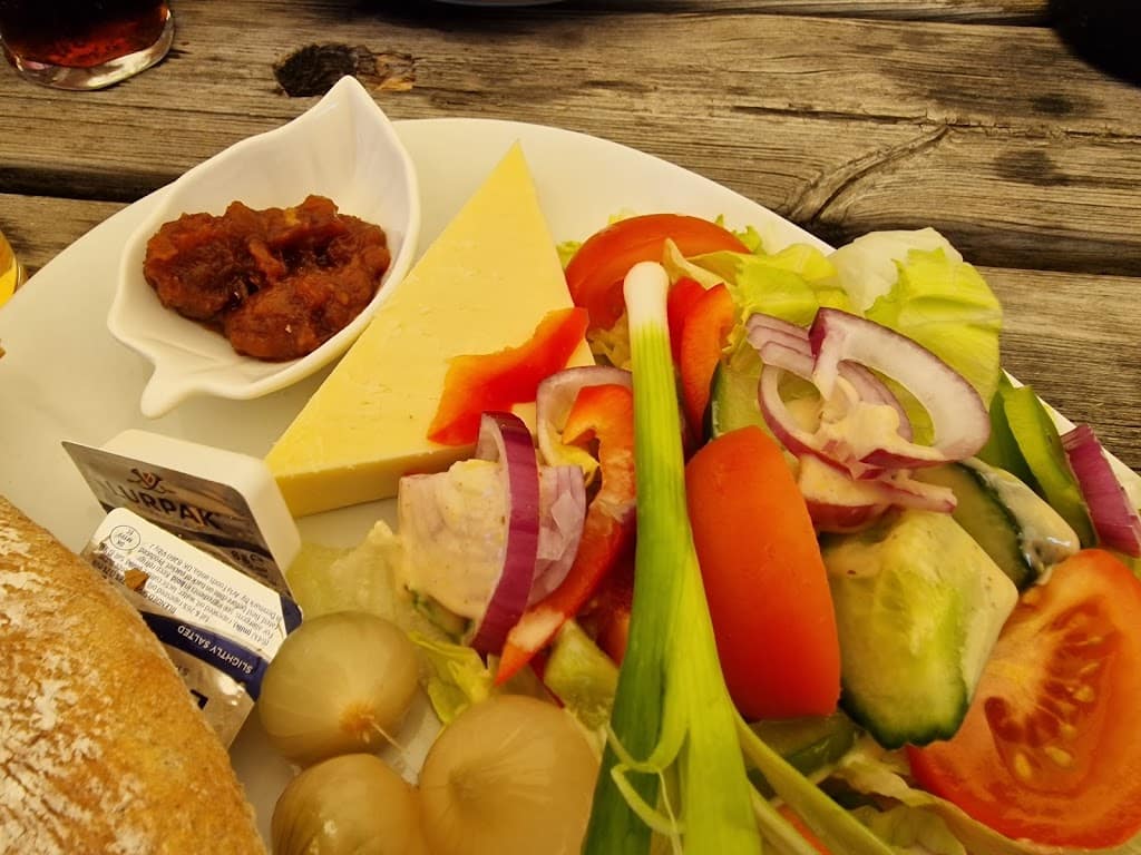 A detailed close-up of a Ploughman's lunch plate. The focus is on a large wedge of cheddar cheese, served alongside a dollop of tangy chutney in a small dish. The colorful salad includes fresh lettuce, thick slices of cucumber, red onion, tomatoes, and red bell peppers. Also on the plate are a few pickled onions, and a small packet of Lurpak butter rests near a slice of rustic bread. The scene is set outdoors on a wooden table, giving a casual, inviting pub garden vibe. The vivid colors of the fresh vegetables contrast with the golden cheese and warm bread, making for an appealing, hearty meal.