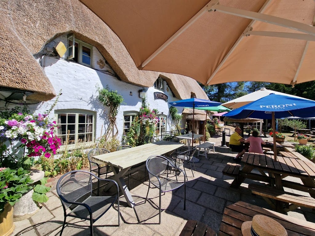 The outdoor patio of the Hatchet Inn features rustic wooden picnic benches and shaded seating under large umbrellas. The beautiful thatched-roof pub is adorned with vibrant hanging flower baskets and climbing plants. A cozy, welcoming atmosphere is enhanced by the quaint whitewashed building and the clear, sunny weather. Ideal for a relaxing meal or drink outdoors, surrounded by greenery and peaceful charm.