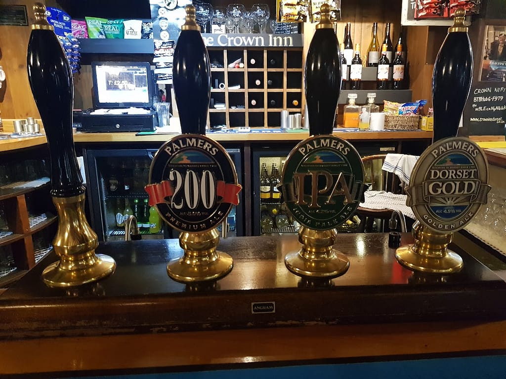 
This image displays a close-up view of the bar at the Crown Inn, showcasing four traditional beer engine handles lined up neatly along the wooden countertop. The handles are used to serve local ales from Palmers Brewery, a well-known brewery in Bridport, Dorset. From left to right, the taps feature:

Palmers 200 Premium Ale: A dark label with "200" in bold red letters on a black background, indicating a premium ale.
Palmers IPA: The green label features the iconic IPA (India Pale Ale) designation.
Palmers Dorset Gold: A golden ale, highlighted with a light-colored label featuring "Dorset Gold."
The backdrop of the bar shows neatly arranged bottles of wine, various bar snacks, and a cash register. The overall atmosphere evokes a traditional, cozy pub ambiance with its wooden accents, making it an inviting spot for visitors to enjoy these local brews.